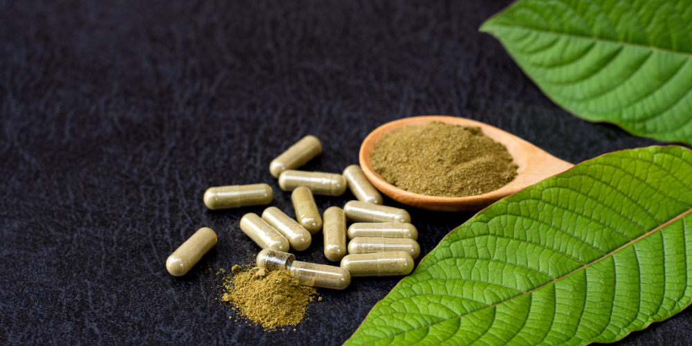 Kratom powder in capsules next to a spoon of kratom powder and a kratom leaf